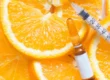 A syringe and a glass vial with a plain white label sit atop sliced oranges in a close-up overhead view, representing Vitamin C Brightening in San Antonio, TX