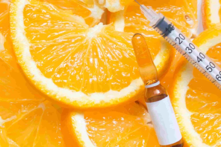 A syringe and a glass vial with a plain white label sit atop sliced oranges in a close-up overhead view, representing Vitamin C Brightening in San Antonio, TX