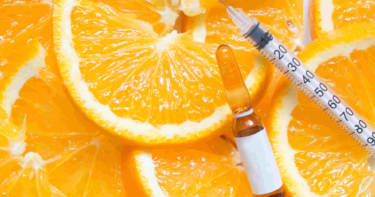 A syringe and a glass vial with a plain white label sit atop sliced oranges in a close-up overhead view, representing Vitamin C Brightening in San Antonio, TX