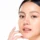 Portrait of a young Asian woman with glowing skin, touching her cheek during Skinvive in San Antonio, TX, against a white background.