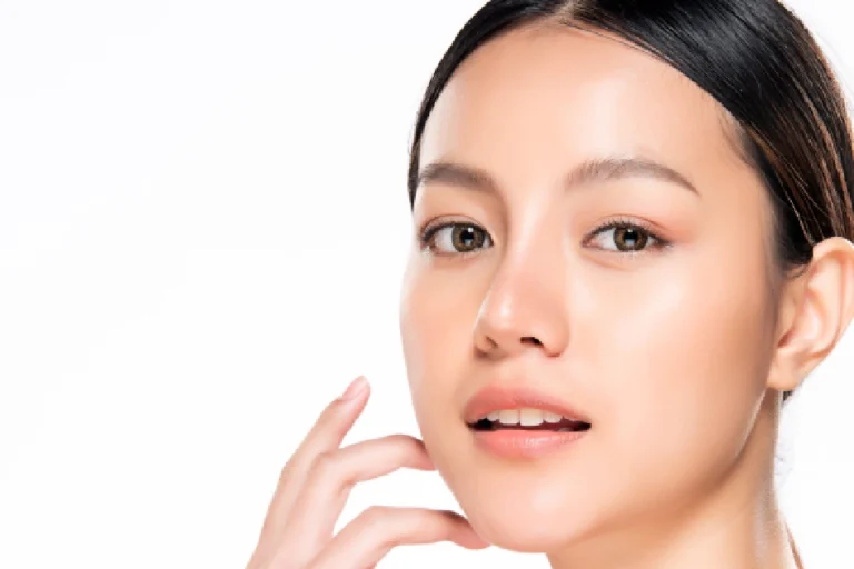 Portrait of a young Asian woman with glowing skin, touching her cheek during Skinvive in San Antonio, TX, against a white background.
