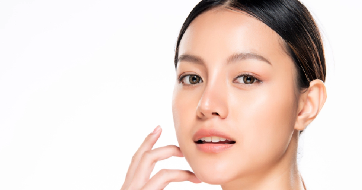 Portrait of a young Asian woman with glowing skin, touching her cheek during Skinvive in San Antonio, TX, against a white background.