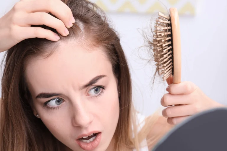 Worried young woman examining hair loss while holding a hairbrush with fallen hair and looking at thinning hairline in the mirror before Hair Restoration in San Antonio.