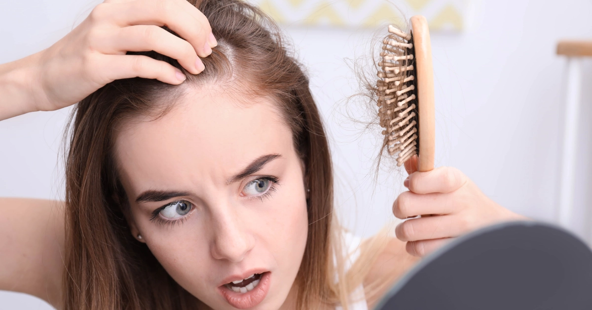 Worried young woman examining hair loss while holding a hairbrush with fallen hair and looking at thinning hairline in the mirror before Hair Restoration in San Antonio.